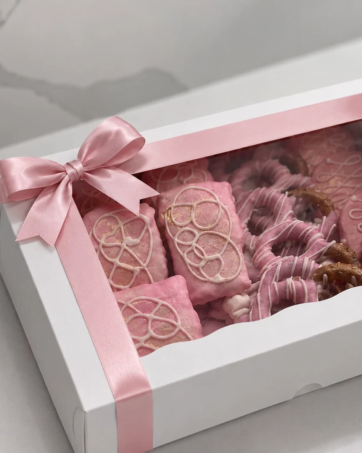 A white gift box tied with a pink ribbon, filled with pink dipped Oreos, pretzels and rice crisp treats